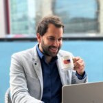 Thibaut Gueant souriant, d'environ 30 ans, aux cheveux bruns et à la barbe, vêtu d'une chemise bleue et d'une veste grise, boit du café dans une tasse blanche ornée du drapeau américain, tout en travaillant sur un ordinateur portable dans un environnement de bureau moderne.