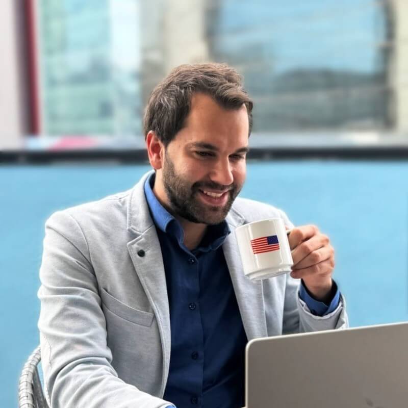 Thibaut Gueant souriant, d'environ 30 ans, aux cheveux bruns et à la barbe, vêtu d'une chemise bleue et d'une veste grise, boit du café dans une tasse blanche ornée du drapeau américain, tout en travaillant sur un ordinateur portable dans un environnement de bureau moderne.